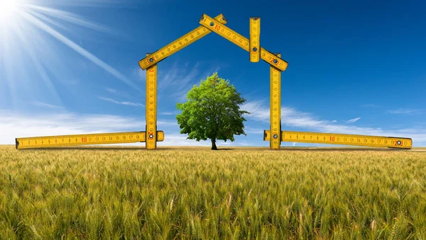 Design or project concept of an ecological house. Folding ruler in the shape of a house, on a wheat field with a green tree. Clear blue sky and sunbeams on background.