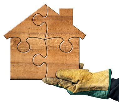 Yapboz parçalarından oluşan küçük ahşap bir evi tutan koruyucu iş eldivenli bir elin yakın çekimi. Beyaz arka planda izole edilmiş. İnşaat endüstrisi konsepti.