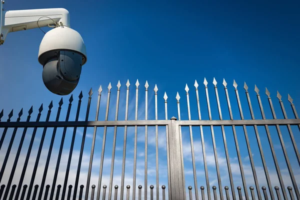 Close-up of a modern security camera monitoring the entrance to a gate of a private property. Wrought iron gate with sharp points on blue sky with clouds and copy space.