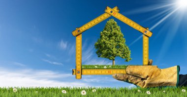 Ekolojik bir evin planlama ve tasarım konsepti. İş eldiveni ile bir evin şeklindeki bir cetveli, çiçekli yeşil bir çayırda ve yeşil bir ağaçla tutarken. Arkaplanda açık mavi gökyüzü.