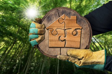 Elleri koruyucu iş eldivenleriyle bir ağaç gövdesinin çapraz kesitini tutarken arka planda yeşil bir orman ve güneş ışınları olan yapboz parçalarından oluşan küçük ahşap bir ev..