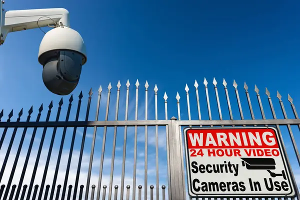 Modern security camera monitoring the entrance to a gate of a private property with a sign with text Warning 24 Hour Video Security Cameras in Use, against a clear blue sky with clouds and copy space.