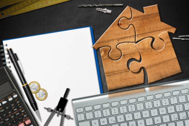 Boş spiral defter, hesap makinesi, Euro bozuklukları, bilgisayar klavyesi, katlama cetveli, kalem ve çalışma araçlarıyla dolu bir masanın üzerindeki yapboz parçalarından yapılmış küçük tahta model ev..