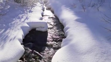 Harika bir kış masalı. Karlı bir orman, bir dağ nehri, dağların üzerinde güneş. Doğa manzarası. Güneş ışınları merceğe güzelce düşer. Kamera aşağıdan yukarıya hareket eder