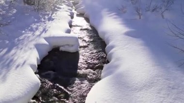 Harika bir kış masalı. Karlı bir orman, bir dağ nehri, dağların üzerinde güneş. Doğa manzarası. Güneş ışınları merceğe güzelce düşer. Kamera aşağıdan yukarıya hareket eder