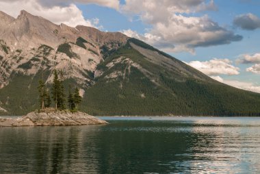 Dağ Gölü manzara panorama insanları - kozalaklı orman - çam ağaçları, manzaralı mavi göl, bulutlu gökyüzü ve ufukta Rocky Dağları. Minnewanka Gölü, Banff il parkı, Alberta, Kanada