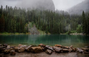 Kanada Rocky Dağları 'nda, Alberta' da, Kanada turizminde çok güzel bir zümrüt yeşili dağ gölü. Yağmurlu bir gün, sis, ağaçların arasından geçen bulutlar. Temiz sakin su, ıslak hava. Ayna Gölü 'nün taşlı kıyıları.