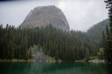 Kanada Rocky Dağları 'nda, Alberta' da, Kanada turizminde çok güzel bir zümrüt yeşili dağ gölü. Yağmurlu bir gün, sis, ağaçların arasından geçen bulutlar. Temiz sakin su, ıslak hava. Ayna Gölü 'nün taşlı kıyıları.