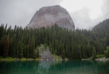 Kanada Rocky Dağları 'nda, Alberta' da, Kanada turizminde çok güzel bir zümrüt yeşili dağ gölü. Yağmurlu bir gün, sis, ağaçların arasından geçen bulutlar. Temiz sakin su, ıslak hava. Ayna Gölü 'nün taşlı kıyıları.