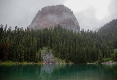 Kanada Rocky Dağları 'nda, Alberta' da, Kanada turizminde çok güzel bir zümrüt yeşili dağ gölü. Yağmurlu bir gün, sis, ağaçların arasından geçen bulutlar. Temiz sakin su, ıslak hava. Ayna Gölü 'nün taşlı kıyıları.