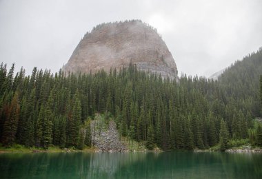 Kanada Rocky Dağları 'nda, Alberta' da, Kanada turizminde çok güzel bir zümrüt yeşili dağ gölü. Yağmurlu bir gün, sis, ağaçların arasından geçen bulutlar. Temiz sakin su, ıslak hava. Ayna Gölü 'nün taşlı kıyıları.