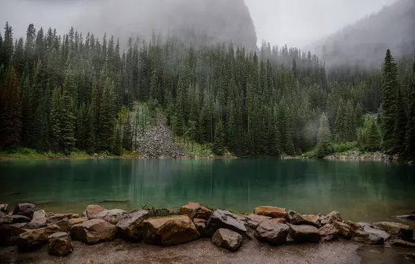 Kanada Rocky Dağları 'nda, Alberta' da, Kanada turizminde çok güzel bir zümrüt yeşili dağ gölü. Yağmurlu bir gün, sis, ağaçların arasından geçen bulutlar. Temiz sakin su, ıslak hava. Ayna Gölü 'nün taşlı kıyıları.