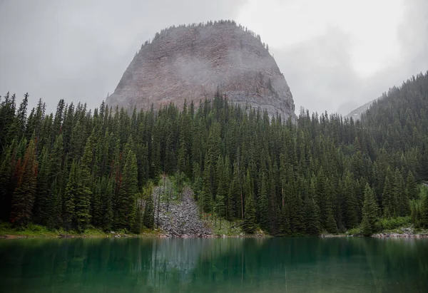 Kanada Rocky Dağları 'nda, Alberta' da, Kanada turizminde çok güzel bir zümrüt yeşili dağ gölü. Yağmurlu bir gün, sis, ağaçların arasından geçen bulutlar. Temiz sakin su, ıslak hava. Ayna Gölü 'nün taşlı kıyıları.