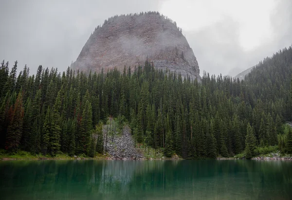 Kanada Rocky Dağları 'nda, Alberta' da, Kanada turizminde çok güzel bir zümrüt yeşili dağ gölü. Yağmurlu bir gün, sis, ağaçların arasından geçen bulutlar. Temiz sakin su, ıslak hava. Ayna Gölü 'nün taşlı kıyıları.