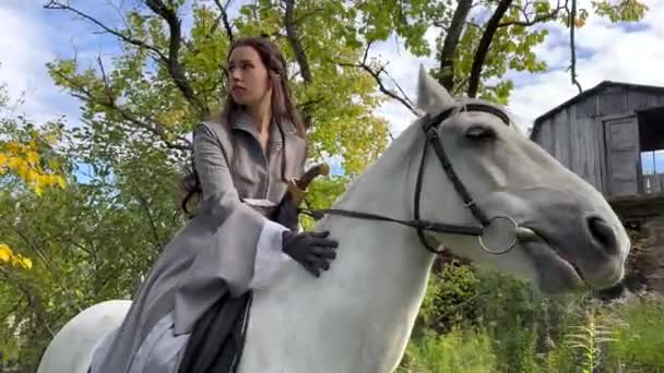 クロークの古代のライダー戦士のイメージの若い女性は白い馬に座って 家や森の背景にそれをストロークします — ストック動画