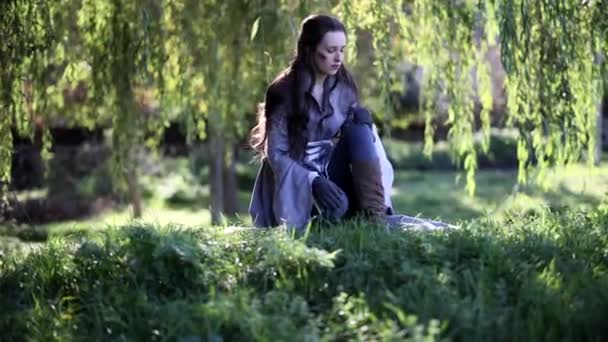 一位年轻女子在古代战士的形象中发现了一个开拓者 她在树下拔草 在森林里看到了它 — 图库视频影像