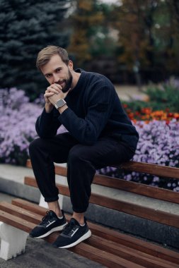 Genç ve mutlu bir adam, çiçek açan yatağın arka planında, parkta bir bankta oturur ve dinlenir..
