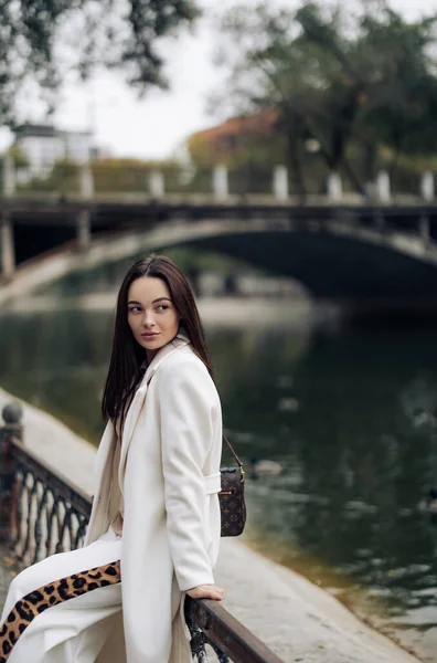 stock image Young pretty woman in white coat with handbag rests in park near lake.