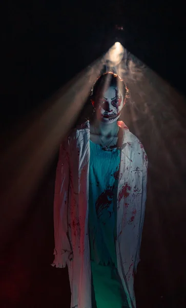 若い女性は顔に恐怖の傷と暗い背景の光の下で流血の服を持つ流血のゾンビを描きます ハロウィンの怖いイメージ — ストック写真
