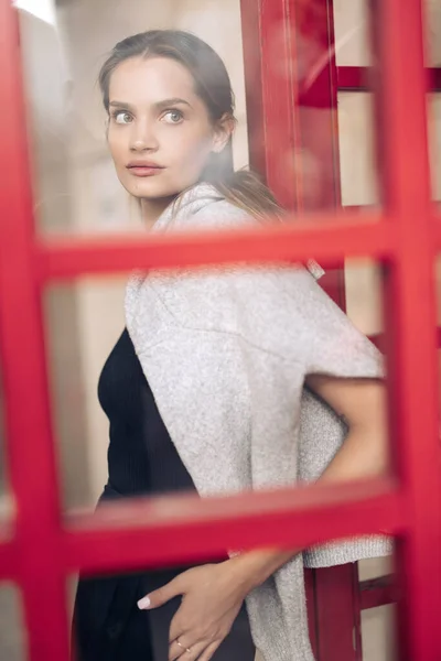 身穿黑色衣服的年轻漂亮女子在大楼的背景下进入红门 穿上衣服透过玻璃看 — 图库照片