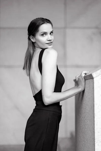 年轻漂亮的女人穿着黑色衣服 光着背站在建筑物附近的街上 穿上衣服侧视图 黑白图像 — 图库照片