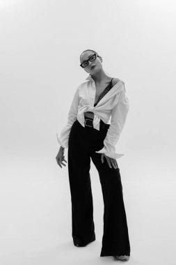 Gözlüklü genç kadın pantolon ve gömlekle beyaz arka planda poz veriyor. Siyah ve beyaz resim.