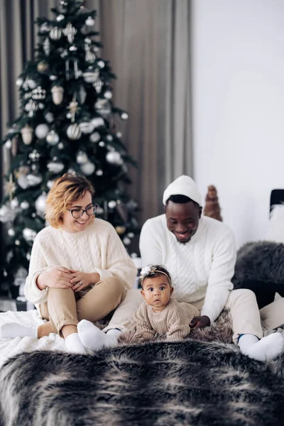 幸せな異人種間の家族はベッドに座って クリスマスツリーを背景に赤ちゃんの娘と遊ぶ 異人種間の家族と団結の概念 — ストック写真