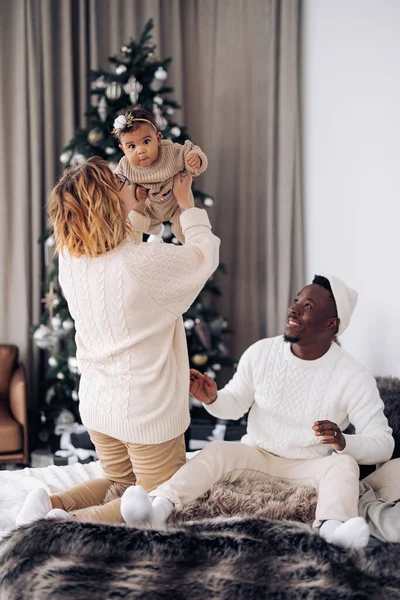 幸せな異人種間の家族は クリスマスツリーを背景に赤ちゃんの娘とベッドで遊ぶ 異人種間の家族と団結の概念 — ストック写真
