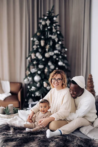 幸せな異人種間の家族はベッドに座って クリスマスツリーを背景に赤ちゃんの娘と遊ぶ 異人種間の家族と団結の概念 — ストック写真