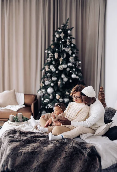 幸せな異人種間の家族はベッドの上に座って クリスマスツリーの背景に赤ちゃんの娘を包含します 異人種間の家族と団結の概念 — ストック写真