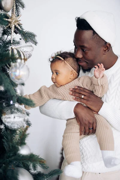 Young african man shows decorations on Christmas tree to his mixed race baby daughter.