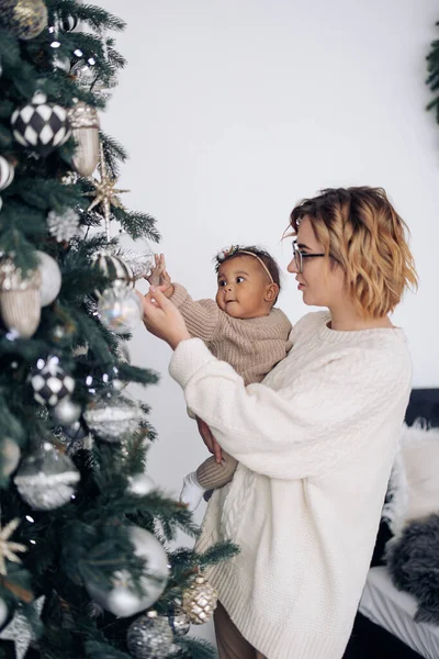 若い白人女性は彼女の混合人種の赤ん坊の娘にクリスマスツリーの装飾を示す 異人種間の家族と団結の概念 — ストック写真