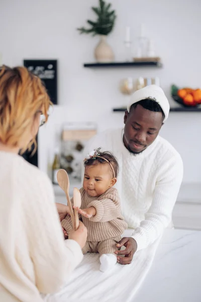 Happy interracial family plays by wooden spoons with their baby daughter on kitchen. Concept of interracial family and unity between different human races.