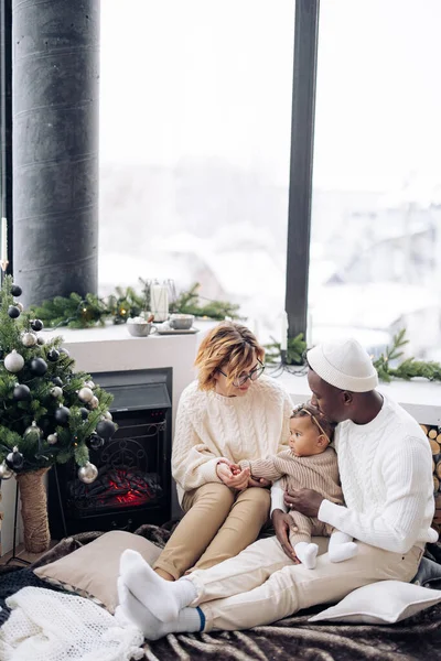 赤ちゃんの娘と幸せな異人種間の家族は クリスマスツリーと装飾を背景に暖炉の近くに座っています 異人種間の家族と団結の概念 — ストック写真