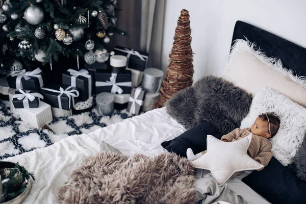 かわいい混合レースの赤ちゃんの女の子は贈り物とクリスマスツリーやボックスを背景に枕の間のベッドの上にあります 最上階だ 異人種間の家族と団結の概念 — ストック写真