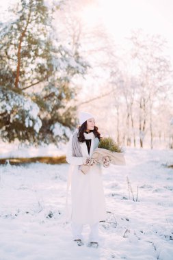 Neşeli genç bir kadın ormanda yürüyor. Güneşli bir kış gününde, karlarla kaplı çam ağaçları arasında. Ellerinde buketle..