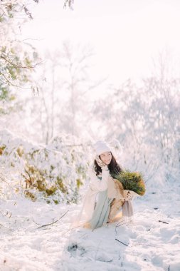 Mutlu genç kadın oturur ve ormanda güler güneşli bir kış gününde ağaçlarla kaplı ağaçların arasında elinde çam dallarından buketlerle..