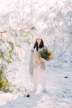 Mutlu genç bir kadın, güneşli bir kış gününde ağaçların arasında elinde çam dallarından buketlerle ormanda yürüyor..