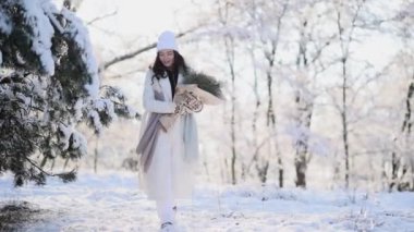 Happy young smiling woman walks in winter forest among snow covered trees with bouquet from pine branches in her hands in sunny day.