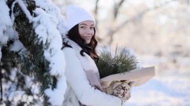 Happy young woman stands and smiles in winter forest near snow covered pine tree in sunny day with bouquet from pine branches in her hands.