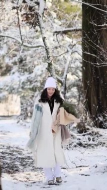 Happy young smiling woman walks in winter forest among snow covered trees in sunny day with bouquet from pine branches in her hands. Vertical video.
