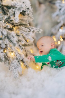 Cute baby boy crawls on floor among artificial snow and touches glowing garlands on Christmas tree.