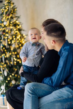 Happy parents play with their baby son against background of Christmas tree with decorations and glowing garlands. Image with bokeh effect.