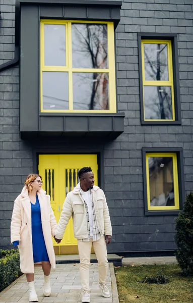 Young interracial couple holds each other hand and walks on street against background of houses. Concept of love relationships and unity between different human races.