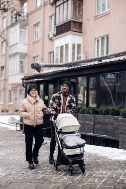 Happy interracial family walk on street and push baby carriage. Concept of interracial family and unity between different human races.