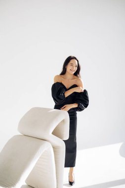 Young woman smiles and poses in studio near armchair in long black dress and high heel shoes against white background.