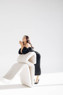 Young woman poses and relaxes with closed eyes in studio near armchair in long black dress and high heel shoes against white background.