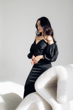 Young woman poses in studio near armchair in black dress against white background.