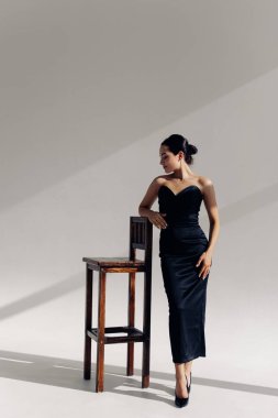 Young woman poses and relaxes with closed eyes in studio standing near chair in long black dress and high heel shoes against white background.
