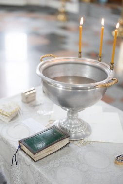 Baptismal font with blessing water and wax candles is on the table covering by white tablecloth near Holy Bible. Newborns baptism rite in Orthodox church.